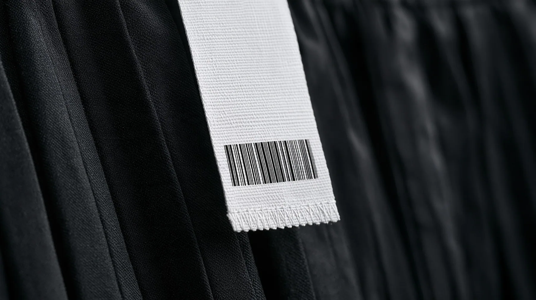 extreme macro photograph of a blank white woven garment tag with a barcode area and a stitched hem hanging from a black pleated coated-nylon skirt, shallow focus, monochrome, the tag face empty for printing, no text,…