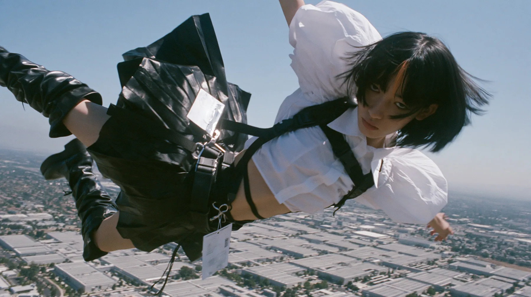 a young woman with a glossy black blunt bob with heavy bangs and one clay-orange streak, a white cropped puff-sleeve shirt, a black pleated skirt with a harness belt and a hanging white garment tag, black knee-high…