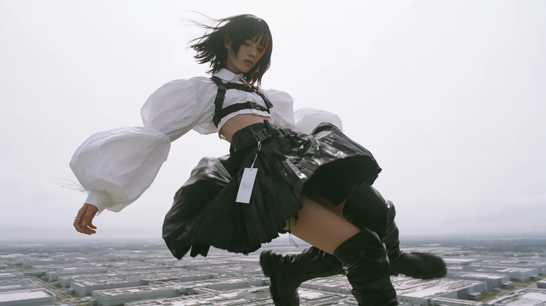 a young woman with a glossy black blunt bob with heavy bangs and one clay-orange streak, a white cropped puff-sleeve shirt, a black pleated skirt with a harness belt and a hanging white garment tag, black knee-high…