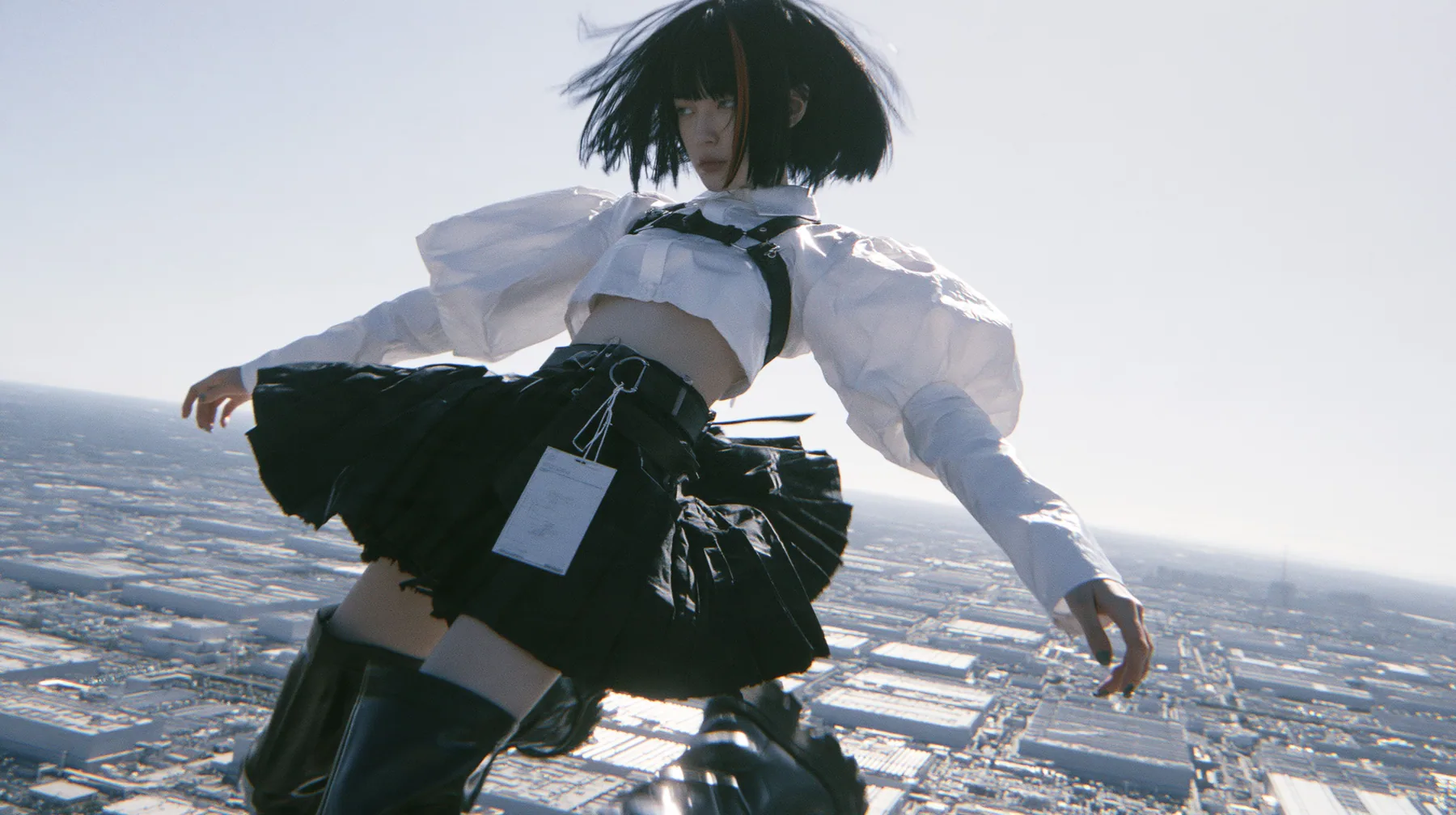 a young woman with a glossy black blunt bob with heavy bangs and one clay-orange streak, a white cropped puff-sleeve shirt, a black pleated skirt with a harness belt and a hanging white garment tag, black knee-high…