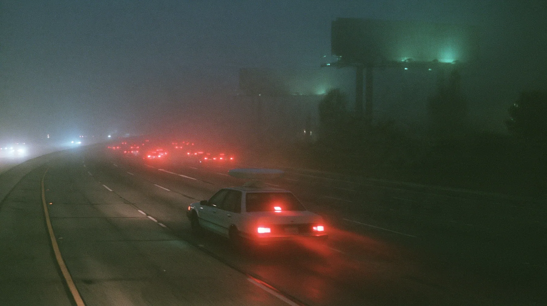 a white driverless robotaxi with a spinning roof sensor driving on the 101 freeway at night in thick fog, huge blank billboards on steel poles fading into the mist above, streams of red tail lights, cinematic 35mm…