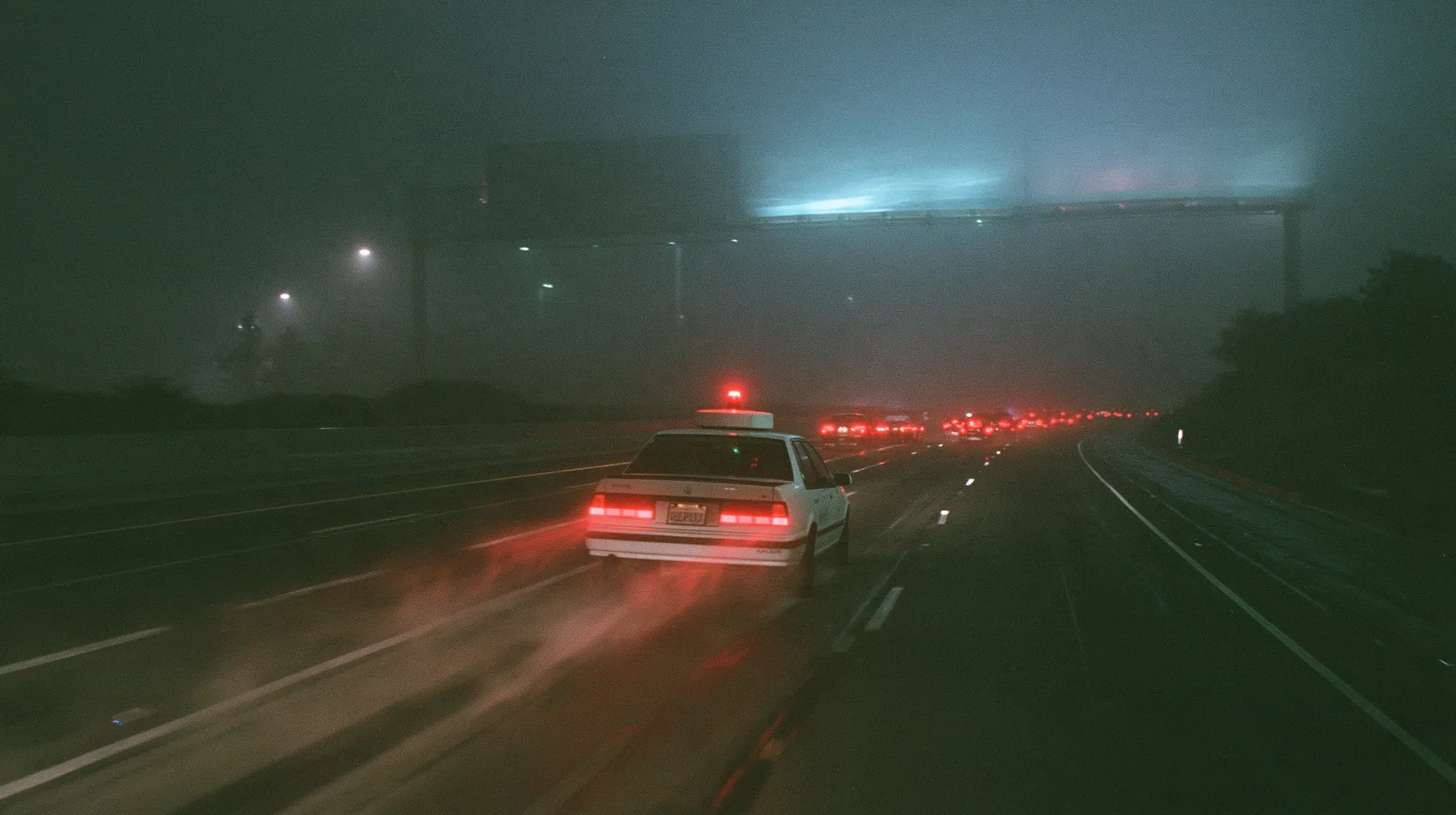 a white driverless robotaxi with a spinning roof sensor driving on the 101 freeway at night in thick fog, huge blank billboards on steel poles fading into the mist above, streams of red tail lights, cinematic 35mm…