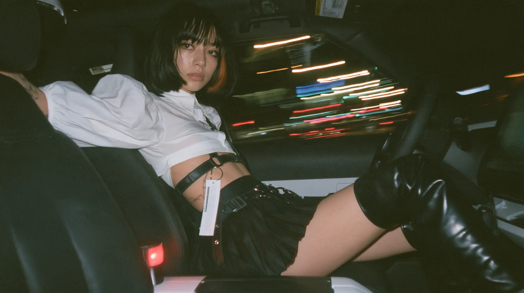 a young woman with a glossy black blunt bob with heavy bangs and one clay-orange streak, a white cropped puff-sleeve shirt, a black pleated skirt with a harness belt and a hanging white garment tag, black knee-high…