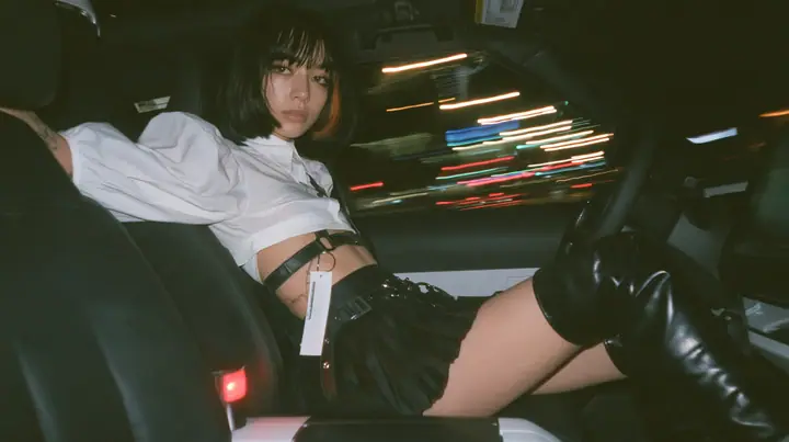 a young woman with a glossy black blunt bob with heavy bangs and one clay-orange streak, a white cropped puff-sleeve shirt, a black pleated skirt with a harness belt and a hanging white garment tag, black knee-high…
