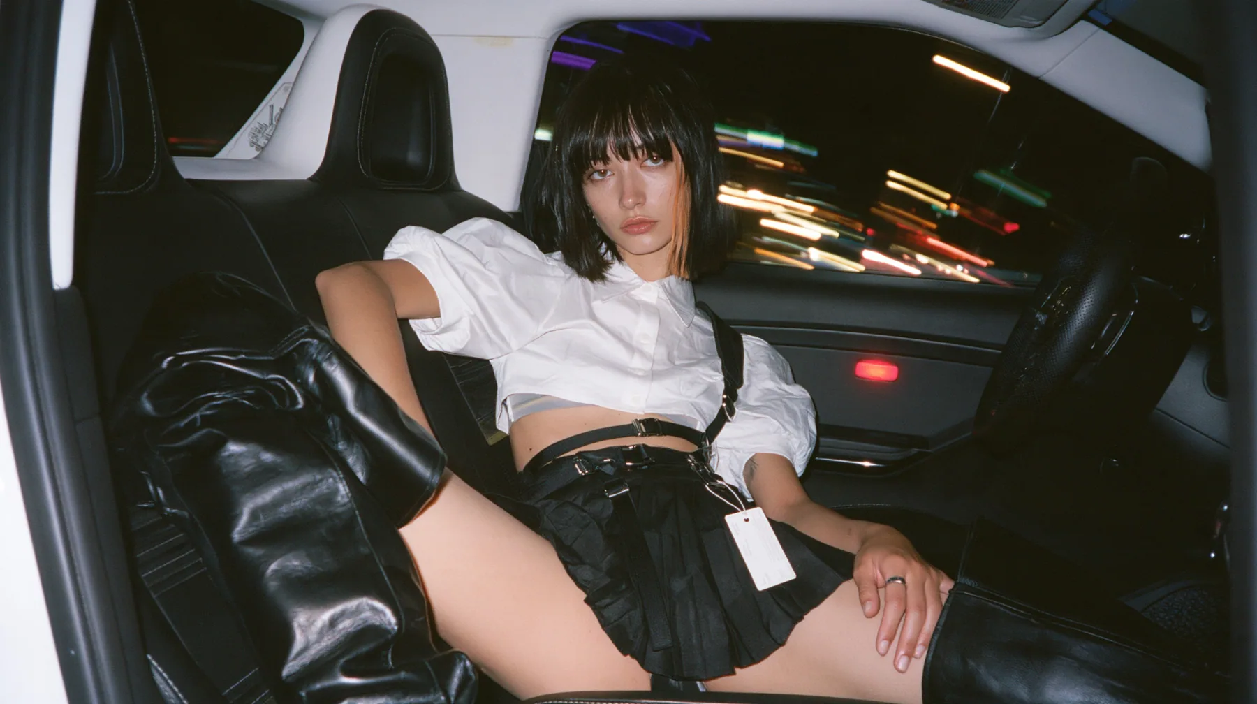 a young woman with a glossy black blunt bob with heavy bangs and one clay-orange streak, a white cropped puff-sleeve shirt, a black pleated skirt with a harness belt and a hanging white garment tag, black knee-high…