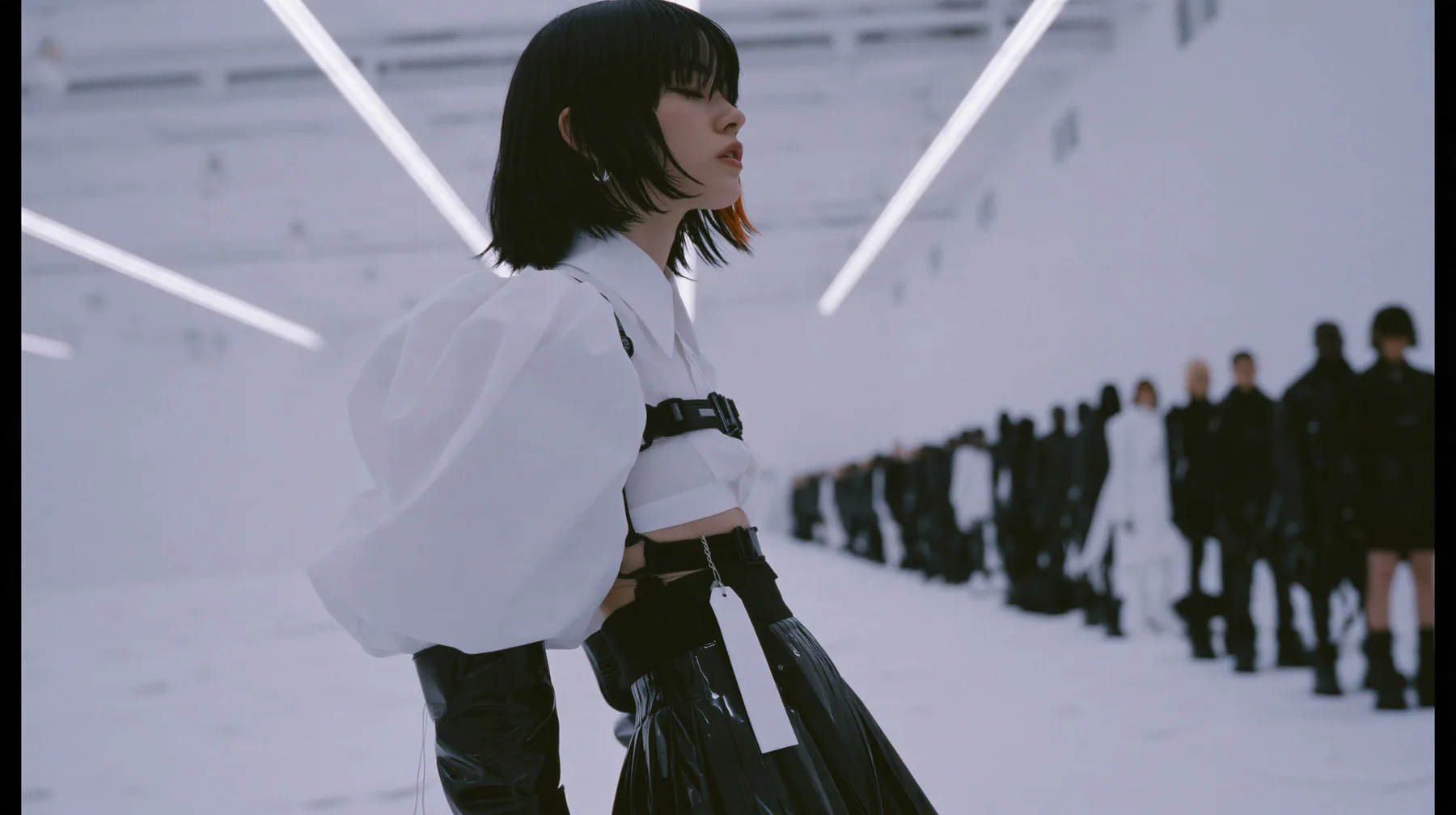 a young woman with a glossy black blunt bob with heavy bangs and one clay-orange streak, a white cropped puff-sleeve shirt, a black pleated skirt with a harness belt and a hanging white garment tag, black knee-high…