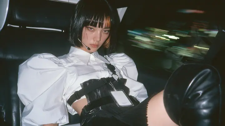 close-up portrait of a young woman with a glossy black blunt bob with heavy bangs and one clay-orange streak, a white cropped puff-sleeve shirt, a black pleated skirt with a harness belt and a hanging white garment…