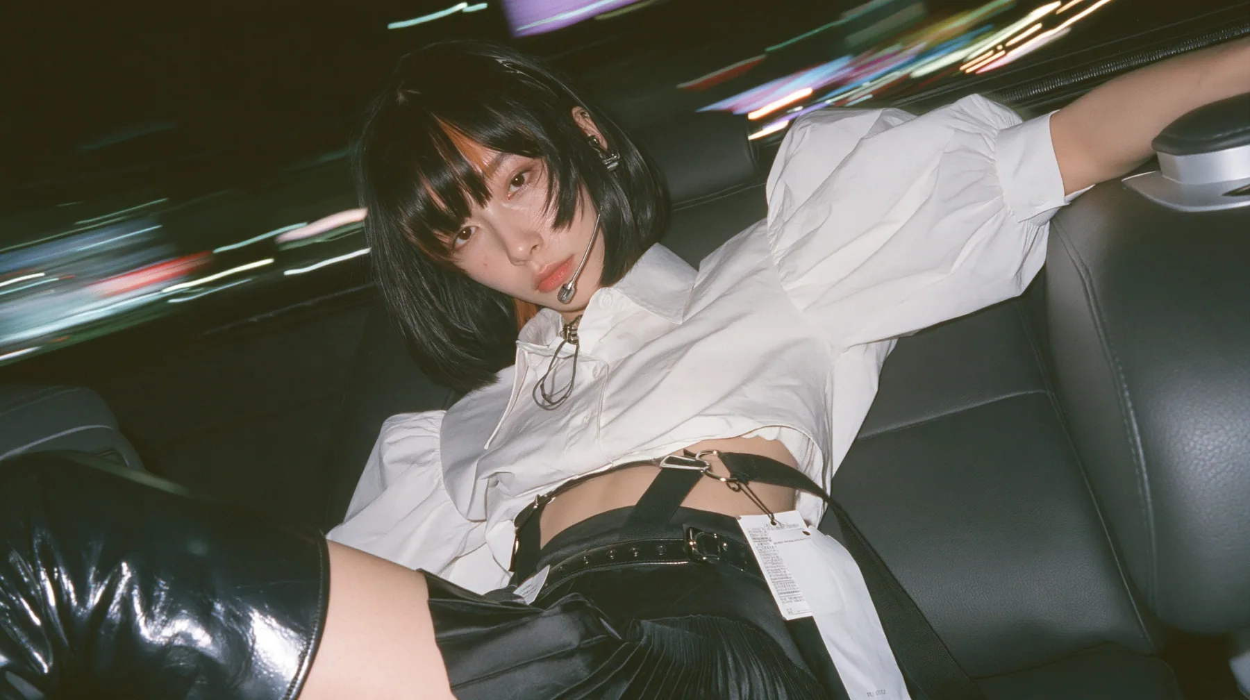 close-up portrait of a young woman with a glossy black blunt bob with heavy bangs and one clay-orange streak, a white cropped puff-sleeve shirt, a black pleated skirt with a harness belt and a hanging white garment…