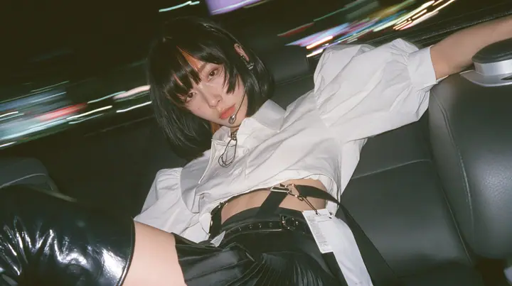 close-up portrait of a young woman with a glossy black blunt bob with heavy bangs and one clay-orange streak, a white cropped puff-sleeve shirt, a black pleated skirt with a harness belt and a hanging white garment…
