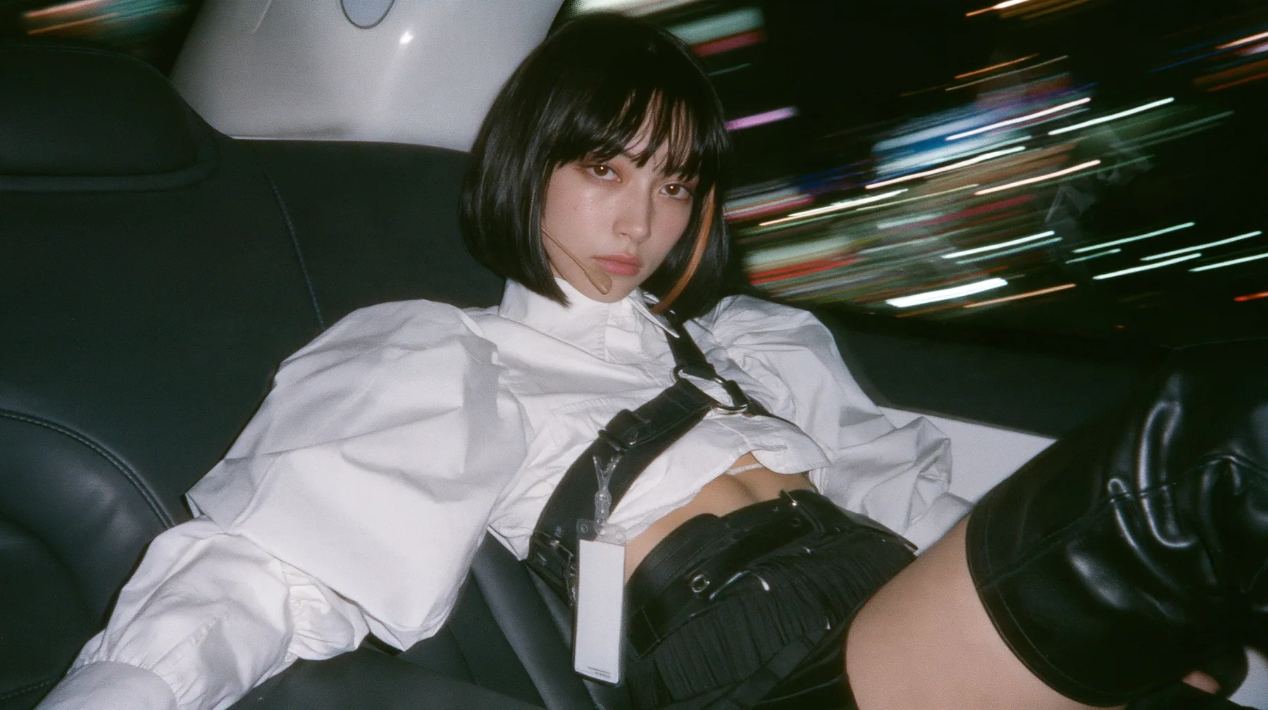 close-up portrait of a young woman with a glossy black blunt bob with heavy bangs and one clay-orange streak, a white cropped puff-sleeve shirt, a black pleated skirt with a harness belt and a hanging white garment…