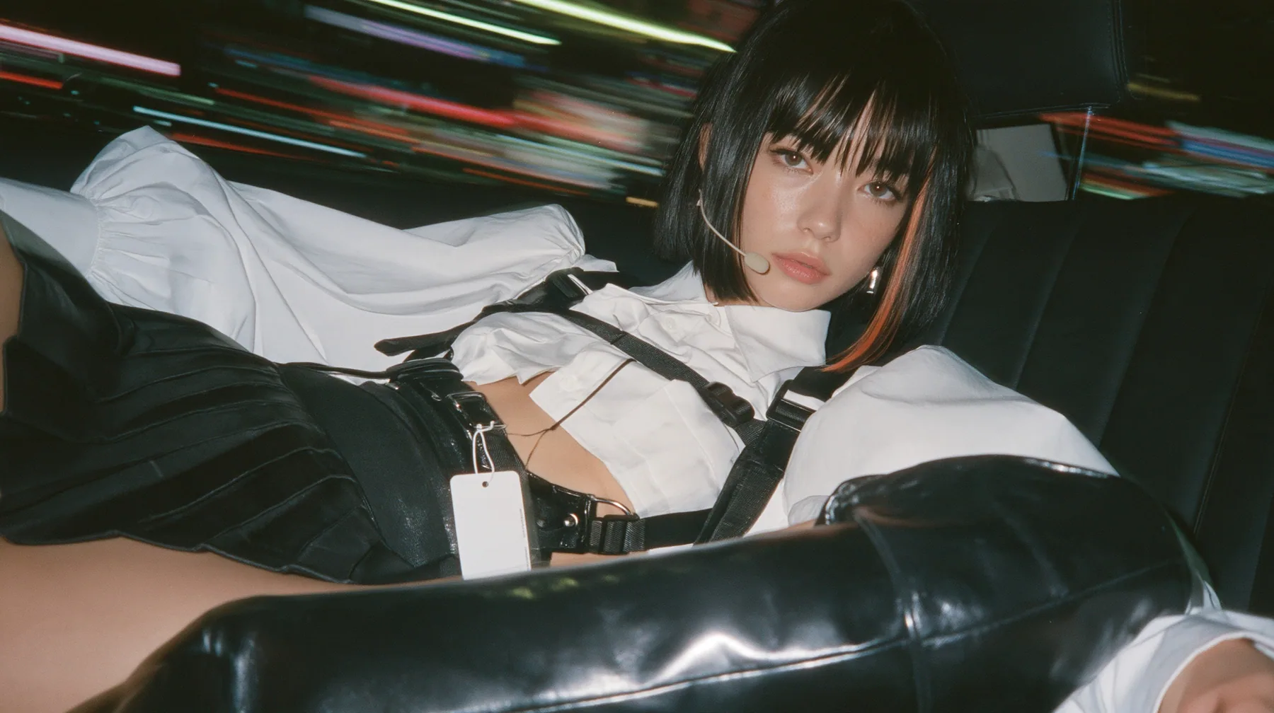 close-up portrait of a young woman with a glossy black blunt bob with heavy bangs and one clay-orange streak, a white cropped puff-sleeve shirt, a black pleated skirt with a harness belt and a hanging white garment…