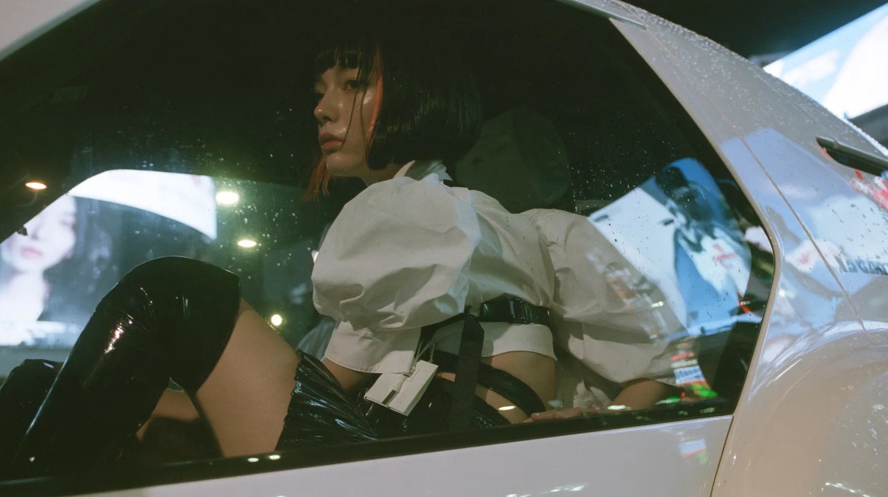 seen from outside through the rear side window of a white driverless robotaxi at night, a young woman with a glossy black blunt bob with heavy bangs and one clay-orange streak, a white cropped puff-sleeve shirt, a…