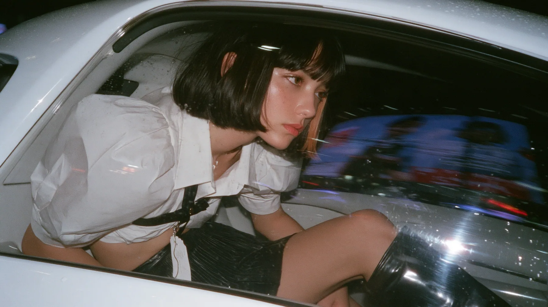 seen from outside through the rear side window of a white driverless robotaxi at night, a young woman with a glossy black blunt bob with heavy bangs and one clay-orange streak, a white cropped puff-sleeve shirt, a…