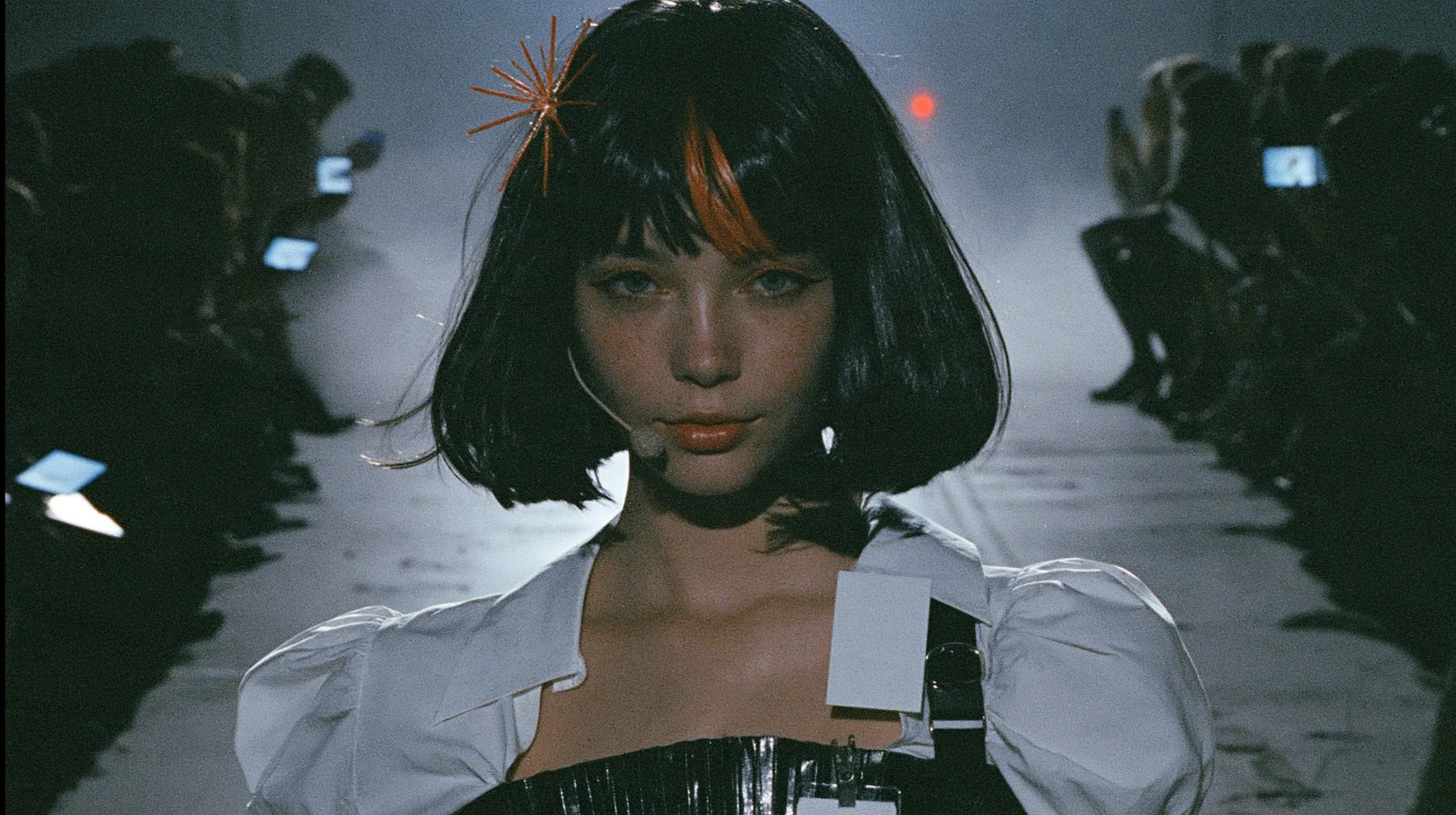 medium shot of a young woman with a glossy black blunt bob with heavy bangs and one clay-orange streak through the bangs, a clay-orange eight-pointed spark hair clip, a thin headset microphone, a white cropped…