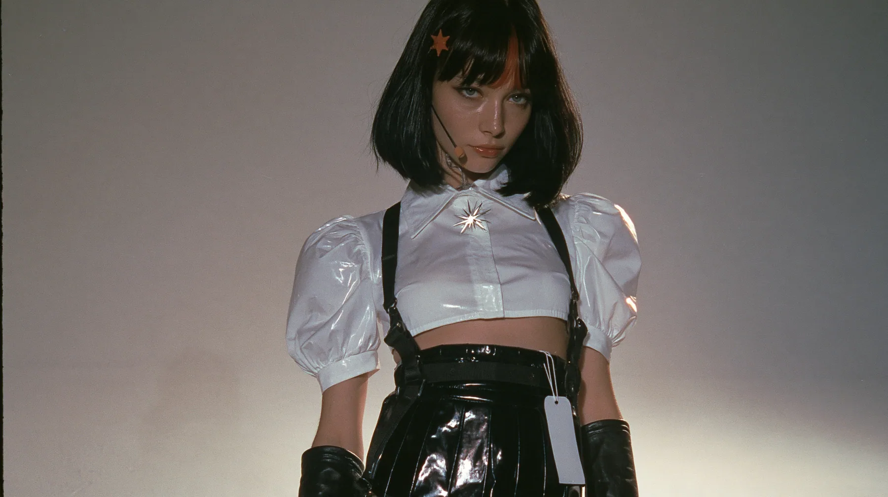 full-length shot of a young woman with a glossy black blunt jaw-length bob with heavy bangs and one clay-orange streak through the bangs, a small flat clay-orange eight-pointed star hair clip, a thin headset…