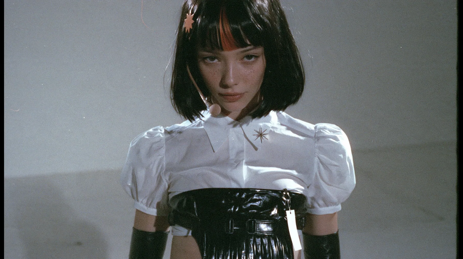 medium shot from the waist up of a young woman with a glossy black blunt jaw-length bob with heavy bangs and one clay-orange streak through the bangs, a small flat clay-orange eight-pointed star hair clip, a thin…