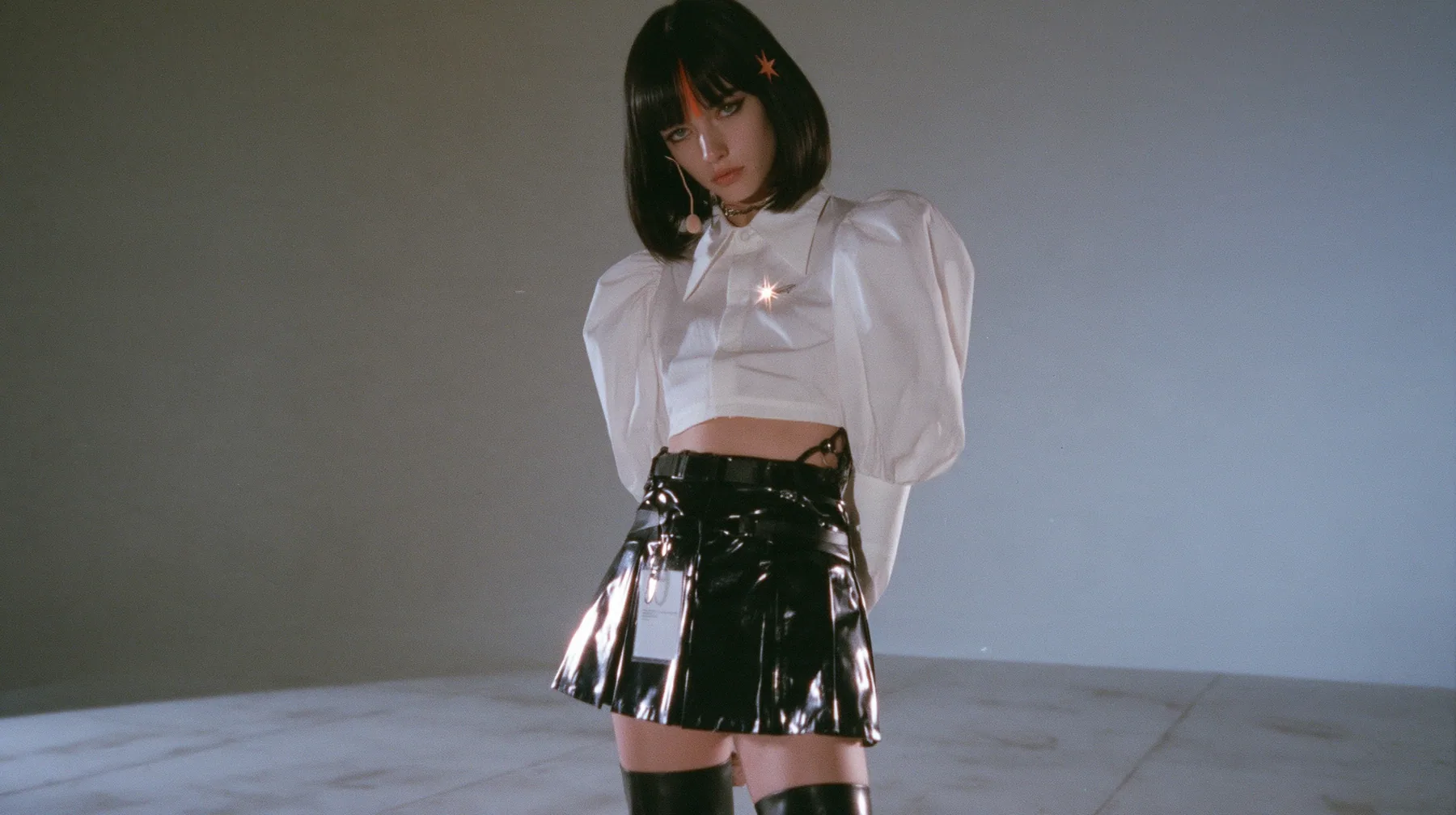 wide shot: a young woman with a glossy black blunt jaw-length bob with heavy bangs and one clay-orange streak through the bangs, a small flat clay-orange eight-pointed star hair clip, a thin headset microphone, a…