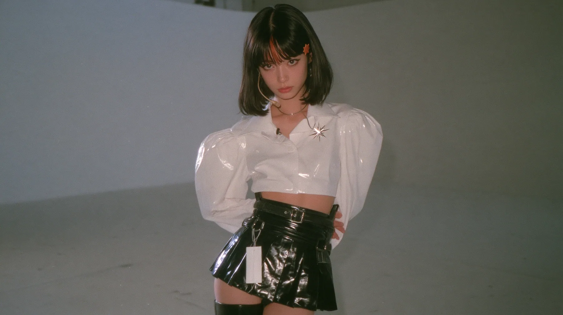 wide shot: a young woman with a glossy black blunt jaw-length bob with heavy bangs and one clay-orange streak through the bangs, a small flat clay-orange eight-pointed star hair clip, a thin headset microphone, a…