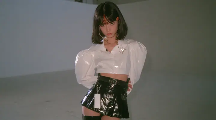 wide shot: a young woman with a glossy black blunt jaw-length bob with heavy bangs and one clay-orange streak through the bangs, a small flat clay-orange eight-pointed star hair clip, a thin headset microphone, a…