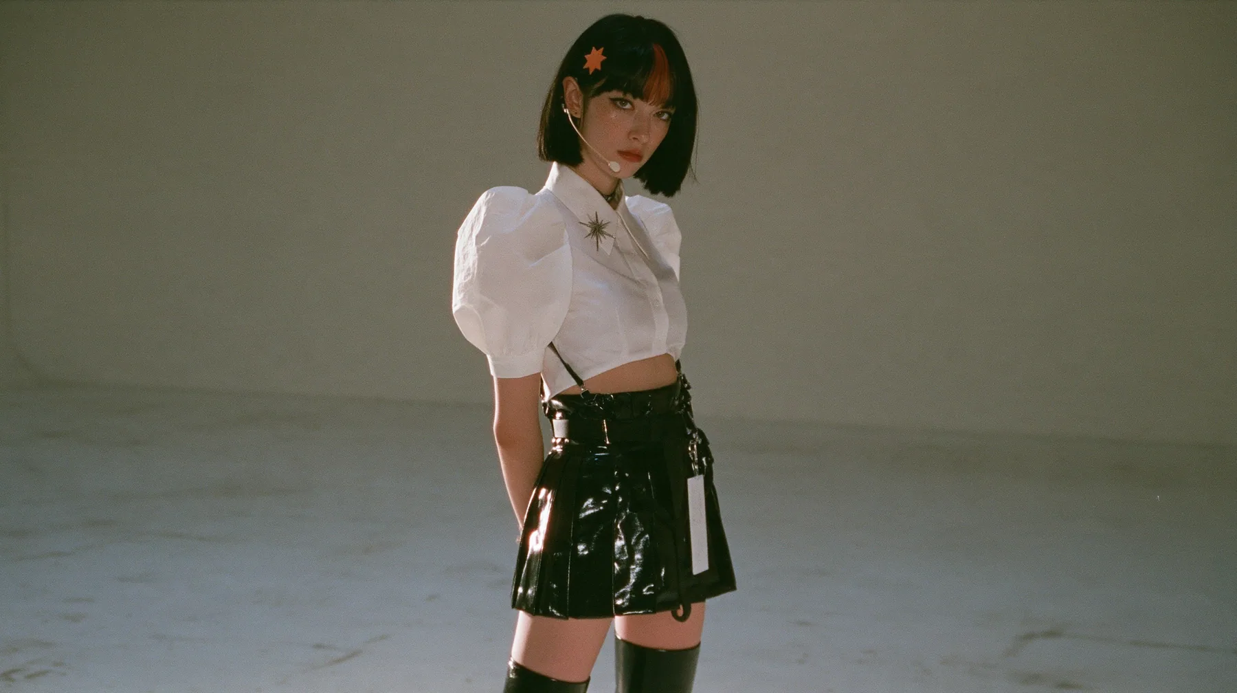 wide shot: a young woman with a glossy black blunt jaw-length bob with heavy bangs and one clay-orange streak through the bangs, a small flat clay-orange eight-pointed star hair clip, a thin headset microphone, a…
