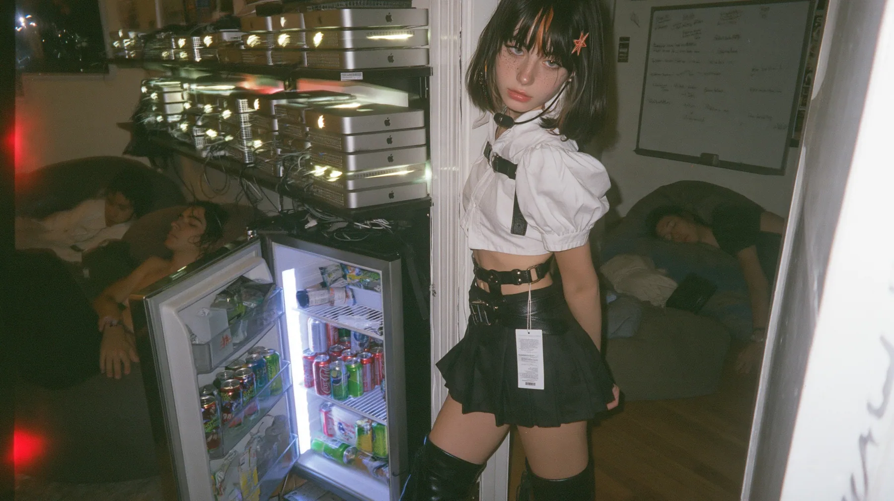 a San Francisco hacker house living room at 3am, stacks of Mac mini computers glowing on shelves and inside an open fridge beside energy drink cans, a whiteboard, people asleep on beanbags, a young Caucasian American…
