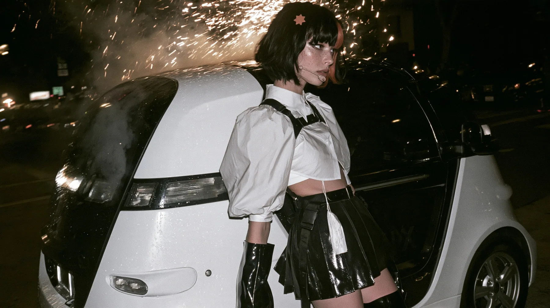 a driverless white robotaxi stopped in a Mission District street at night while fireworks burst low overhead and sparks rain onto its roof, smoke, a young Caucasian American woman with pale skin and light freckles, a…