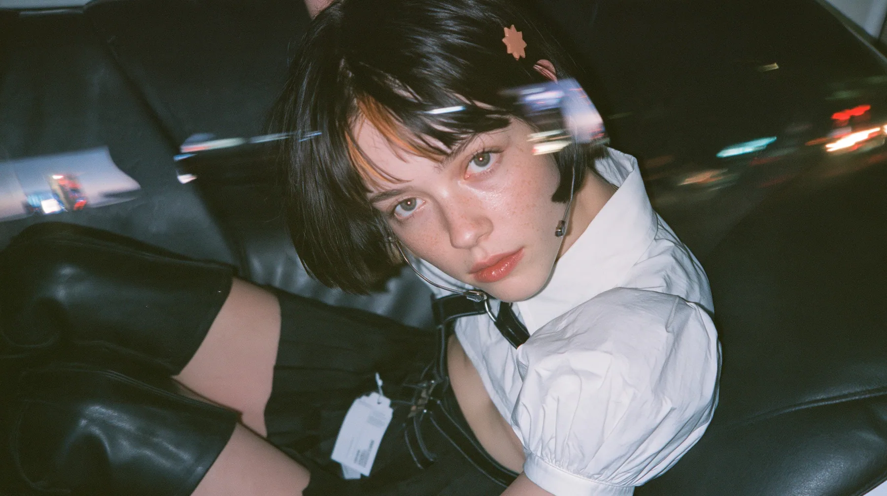 close-up portrait of a young Caucasian American woman with pale skin and light freckles, a glossy black blunt jaw-length bob with heavy bangs and one clay-orange streak through the bangs, a small flat clay-orange…