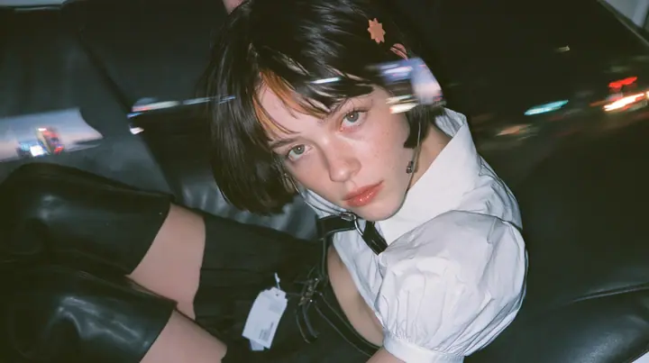 close-up portrait of a young Caucasian American woman with pale skin and light freckles, a glossy black blunt jaw-length bob with heavy bangs and one clay-orange streak through the bangs, a small flat clay-orange…
