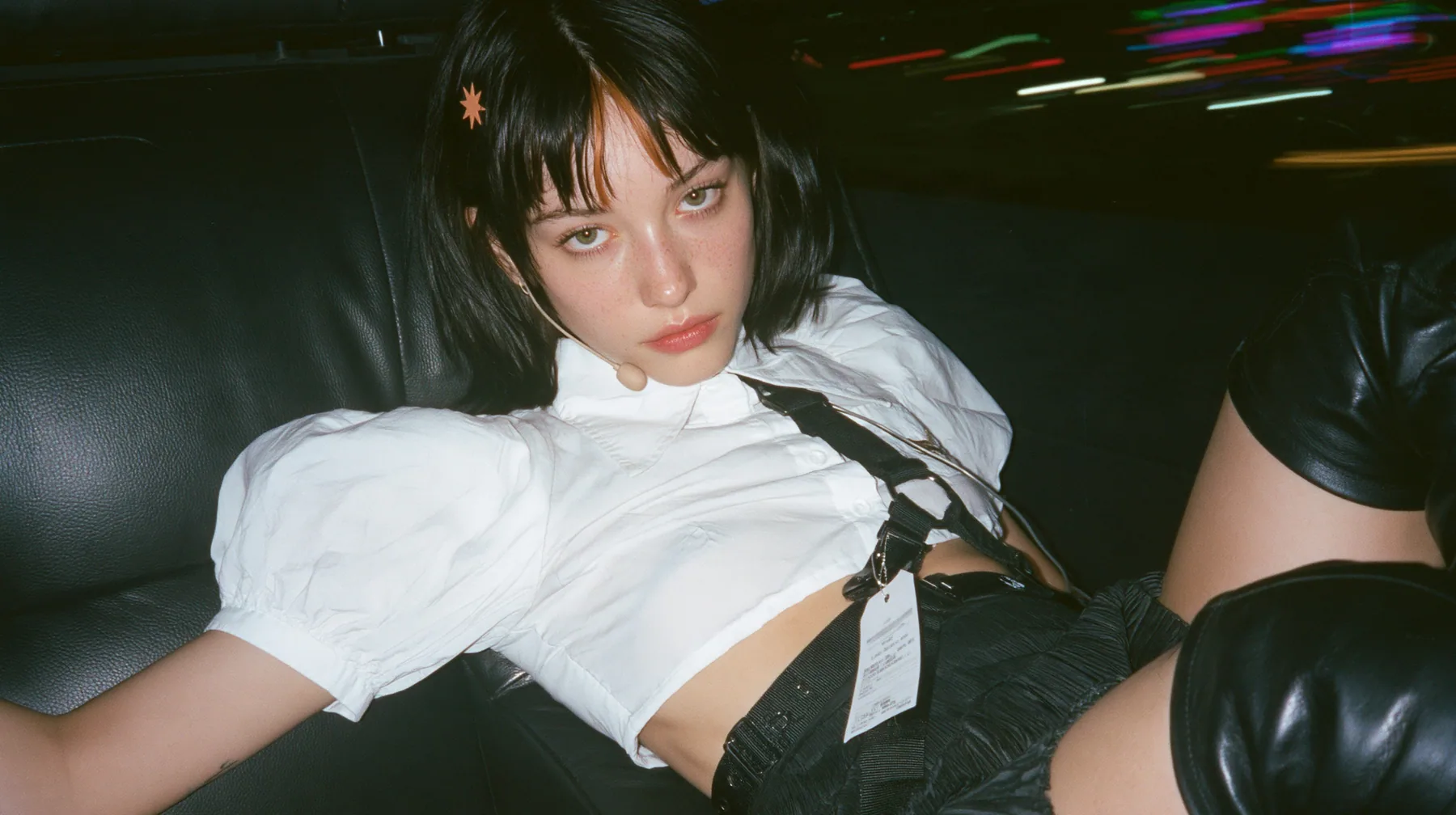 close-up portrait of a young Caucasian American woman with pale skin and light freckles, a glossy black blunt jaw-length bob with heavy bangs and one clay-orange streak through the bangs, a small flat clay-orange…