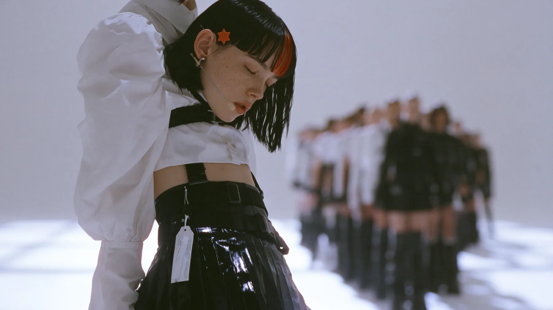 a young Caucasian American woman with pale skin and light freckles, a glossy black blunt jaw-length bob with heavy bangs and one clay-orange streak through the bangs, a small flat clay-orange eight-pointed star hair…