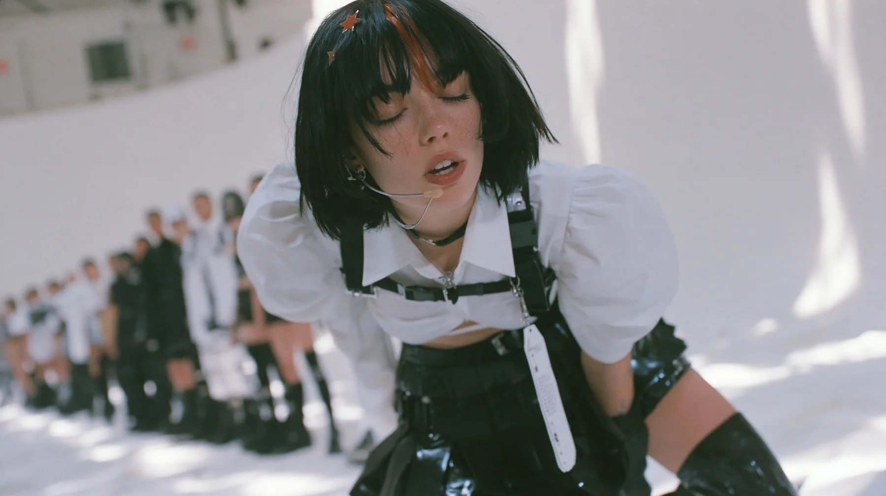 a young Caucasian American woman with pale skin and light freckles, a glossy black blunt jaw-length bob with heavy bangs and one clay-orange streak through the bangs, a small flat clay-orange eight-pointed star hair…