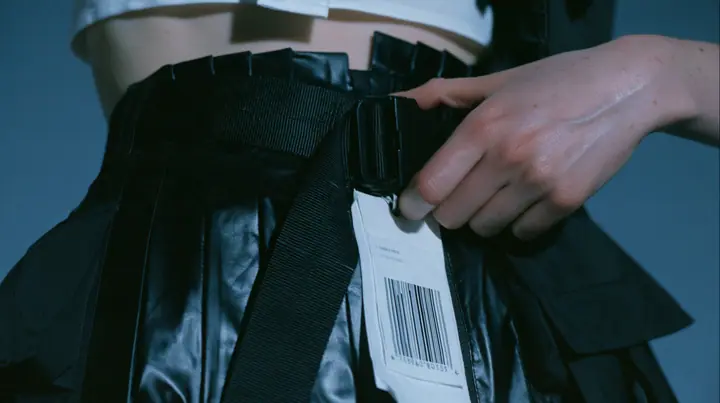 close-up of a young Caucasian American woman with pale skin and light freckles,'s hand pulling a black utility harness strap tight over a black pleated coated-nylon skirt, a white garment tag with a barcode hanging…
