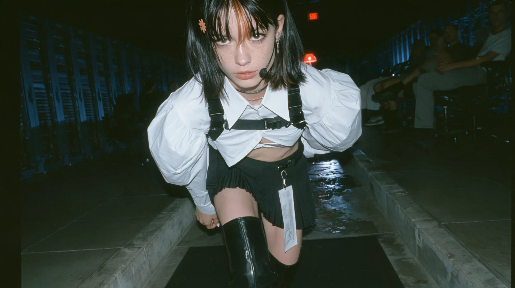 low-angle full-length view of a young Caucasian American woman with pale skin and light freckles, a glossy black blunt jaw-length bob with heavy bangs and one clay-orange streak through the bangs, a small flat…