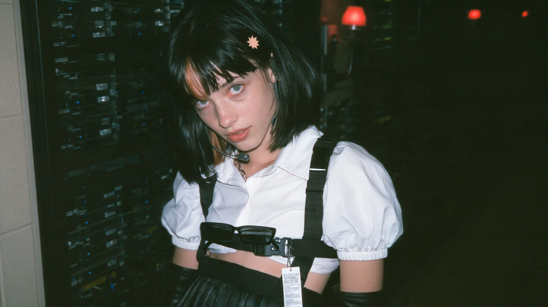 medium close-up of a young Caucasian American woman with pale skin and light freckles, a glossy black blunt jaw-length bob with heavy bangs and one clay-orange streak through the bangs, a small flat clay-orange…