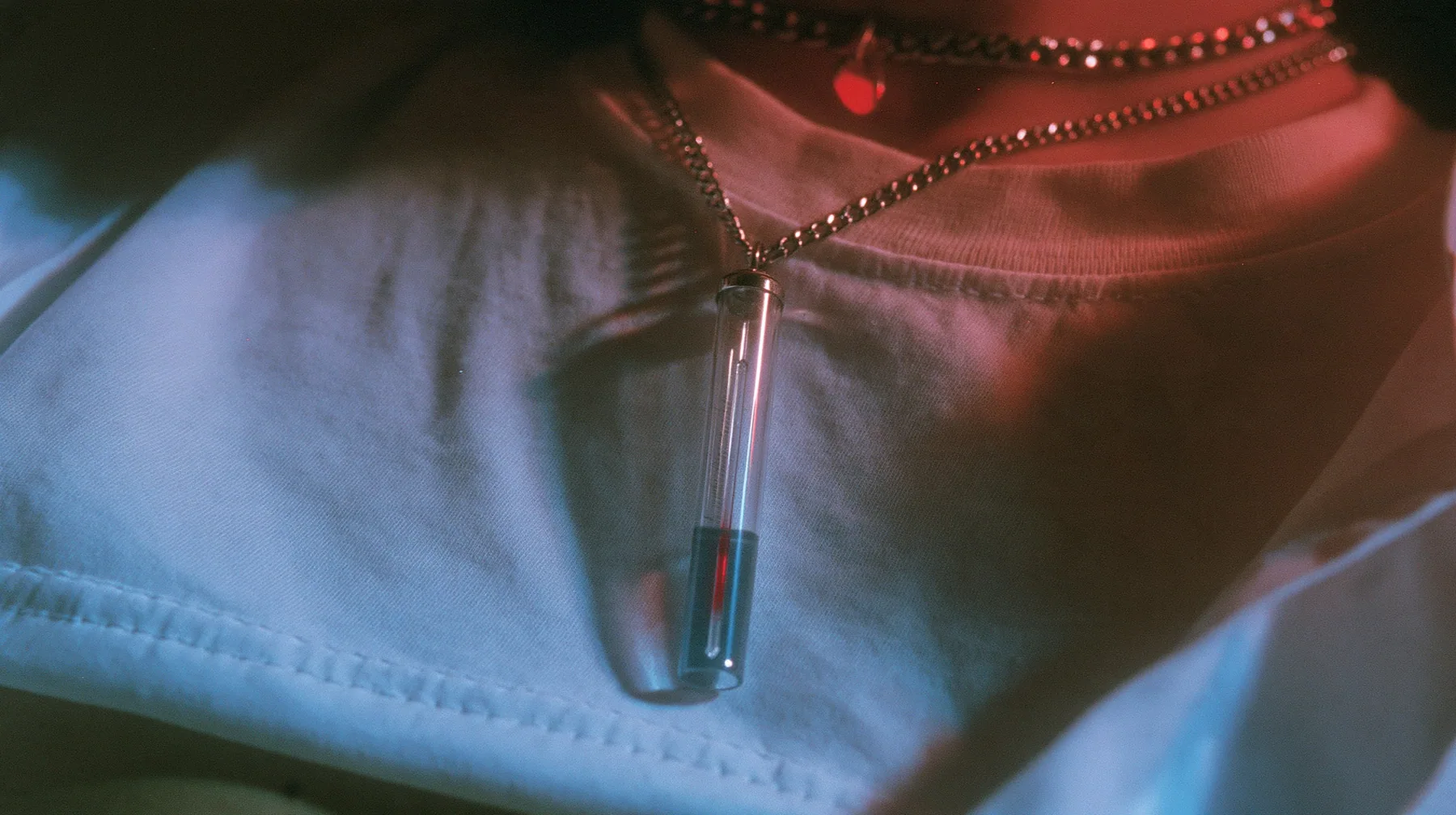 extreme close-up of a slim glass thermometer pendant on a long steel chain resting on a white cropped shirt, the mercury halfway, dark background, the right half empty, blue-black #0f1216 and steel blue, one small…