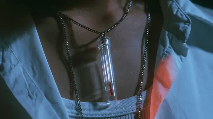 extreme close-up of a slim glass thermometer pendant on a long steel chain resting on a white cropped shirt, the mercury halfway, dark background, the right half empty, blue-black #0f1216 and steel blue, one small…