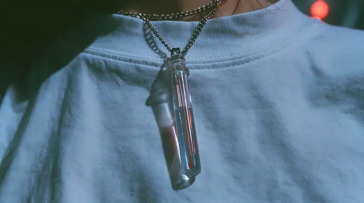 extreme close-up of a slim glass thermometer pendant on a long steel chain resting on a white cropped shirt, the mercury halfway, dark background, the right half empty, blue-black #0f1216 and steel blue, one small…