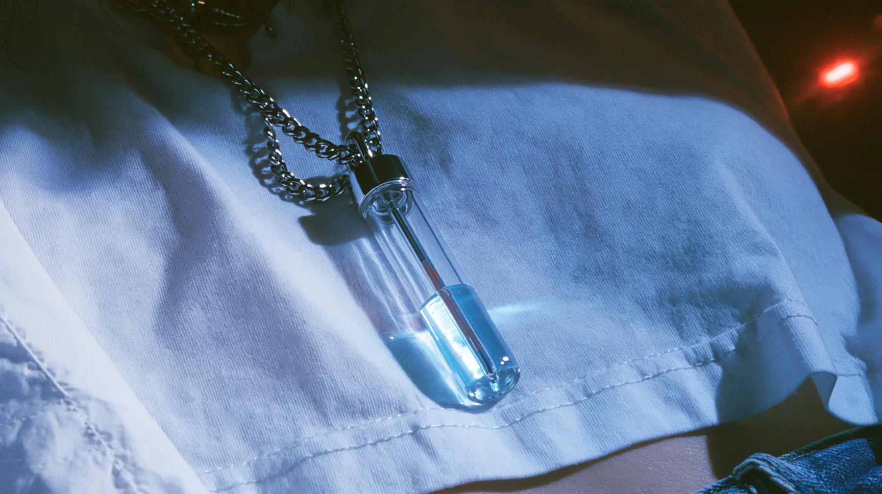 extreme close-up of a slim glass thermometer pendant on a long steel chain resting on a white cropped shirt, the mercury halfway, dark background, the right half empty, blue-black #0f1216 and steel blue, one small…
