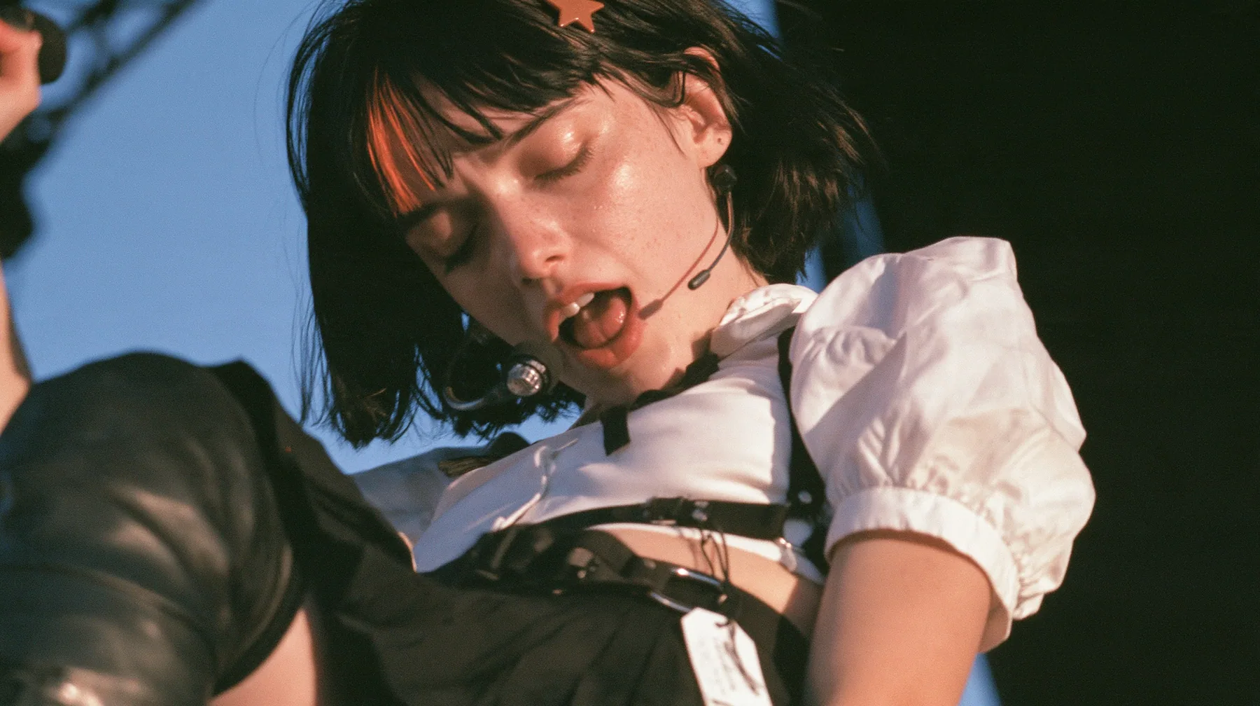 close-up of a young Caucasian American woman with pale skin and light freckles, a glossy black blunt jaw-length bob with heavy bangs and one clay-orange streak through the bangs, a small flat clay-orange…