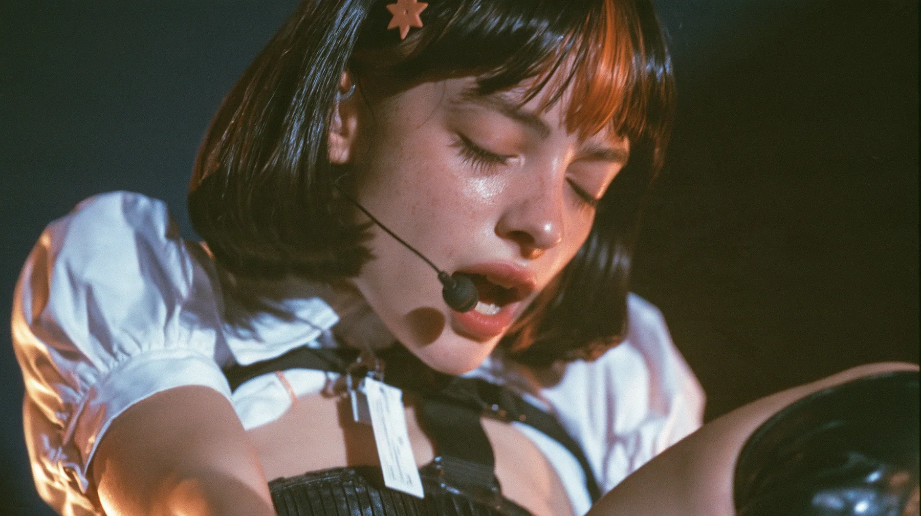 close-up of a young Caucasian American woman with pale skin and light freckles, a glossy black blunt jaw-length bob with heavy bangs and one clay-orange streak through the bangs, a small flat clay-orange…