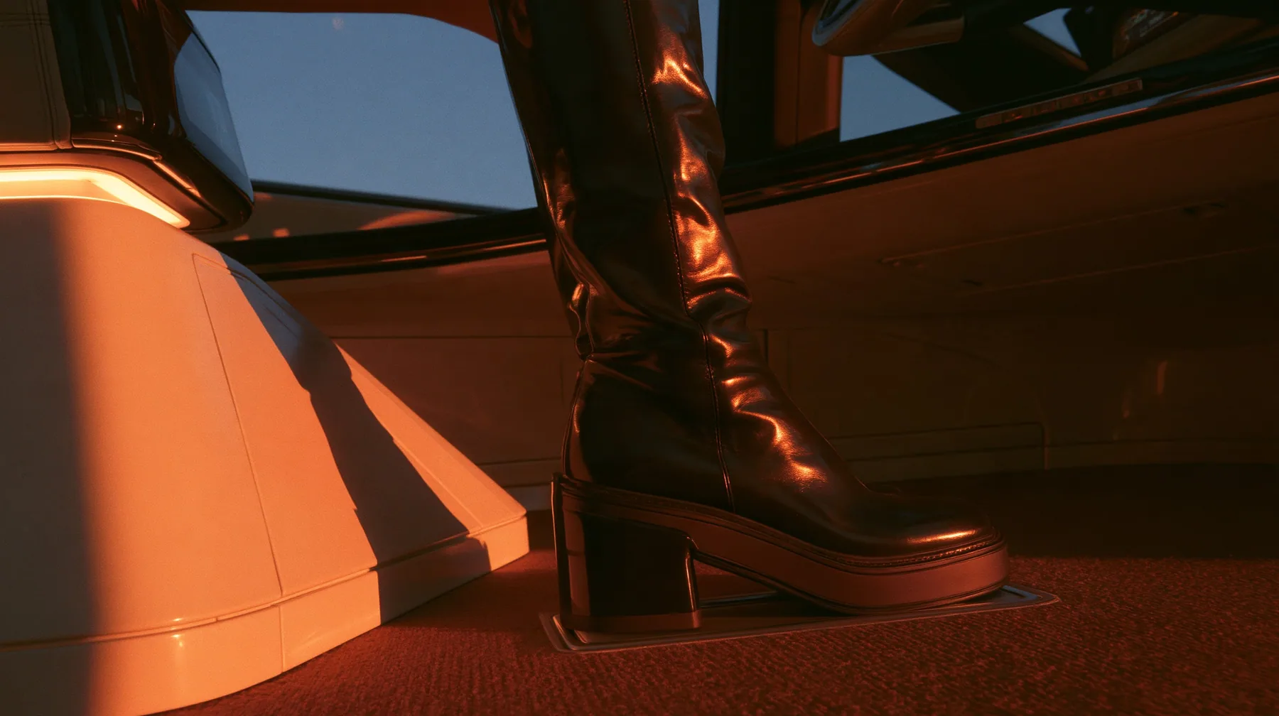 close-up of a black knee-high boot pressing flat on the bare floor of a driverless robotaxi, no pedals, no steering wheel, night light sweeping across, blue-black night turning to first light, clay-orange #D97757…