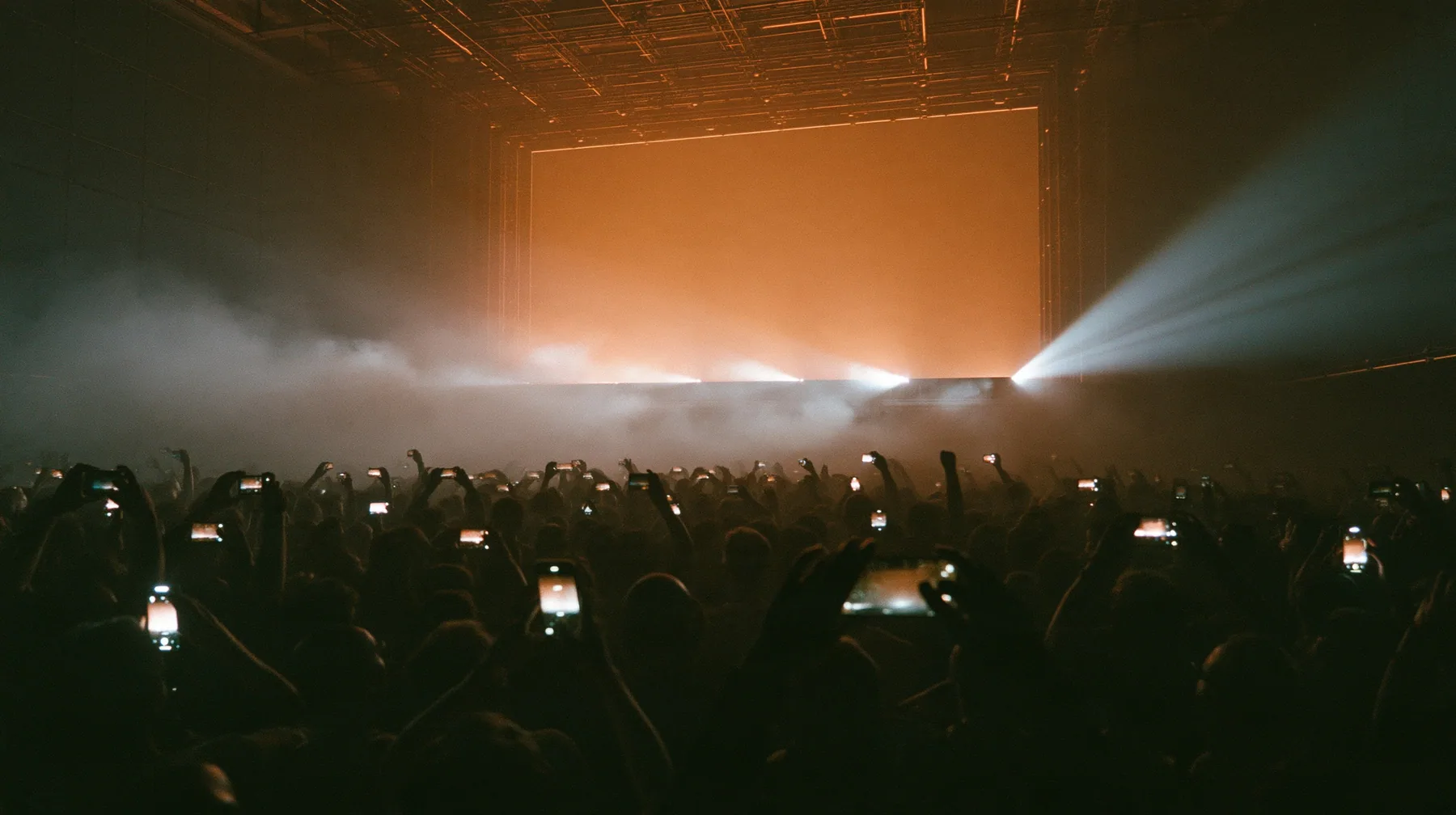a dark crowd of front-row spectators raising glowing phones above their heads in a data hall, backlit fog, warm light from a giant board behind, blue-black night turning to first light, clay-orange #D97757 light as…