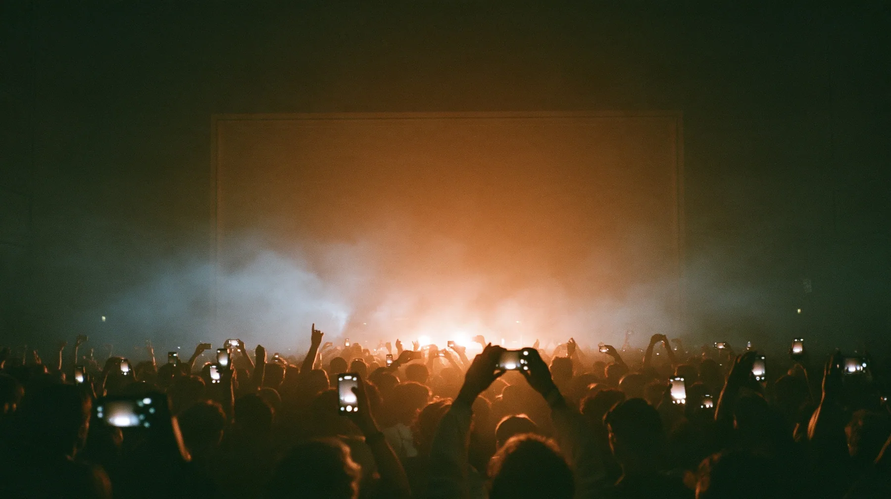 a dark crowd of front-row spectators raising glowing phones above their heads in a data hall, backlit fog, warm light from a giant board behind, blue-black night turning to first light, clay-orange #D97757 light as…