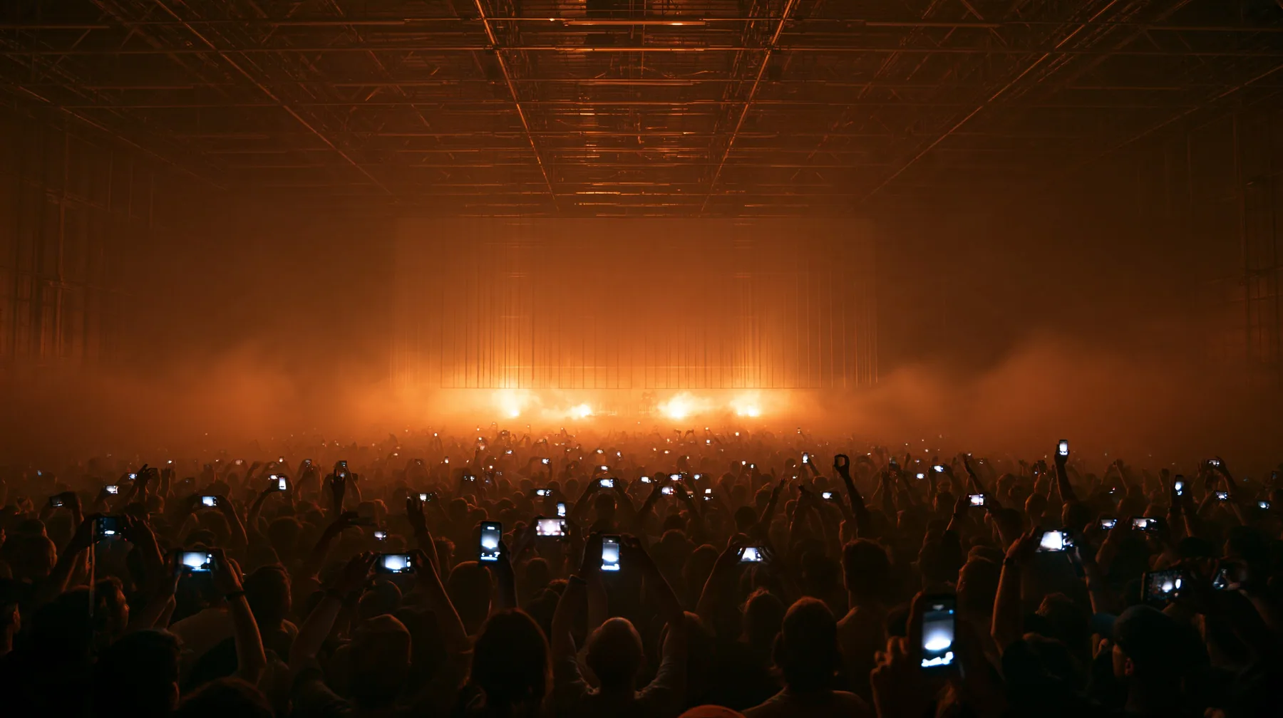 a dark crowd of front-row spectators raising glowing phones above their heads in a data hall, backlit fog, warm light from a giant board behind, blue-black night turning to first light, clay-orange #D97757 light as…