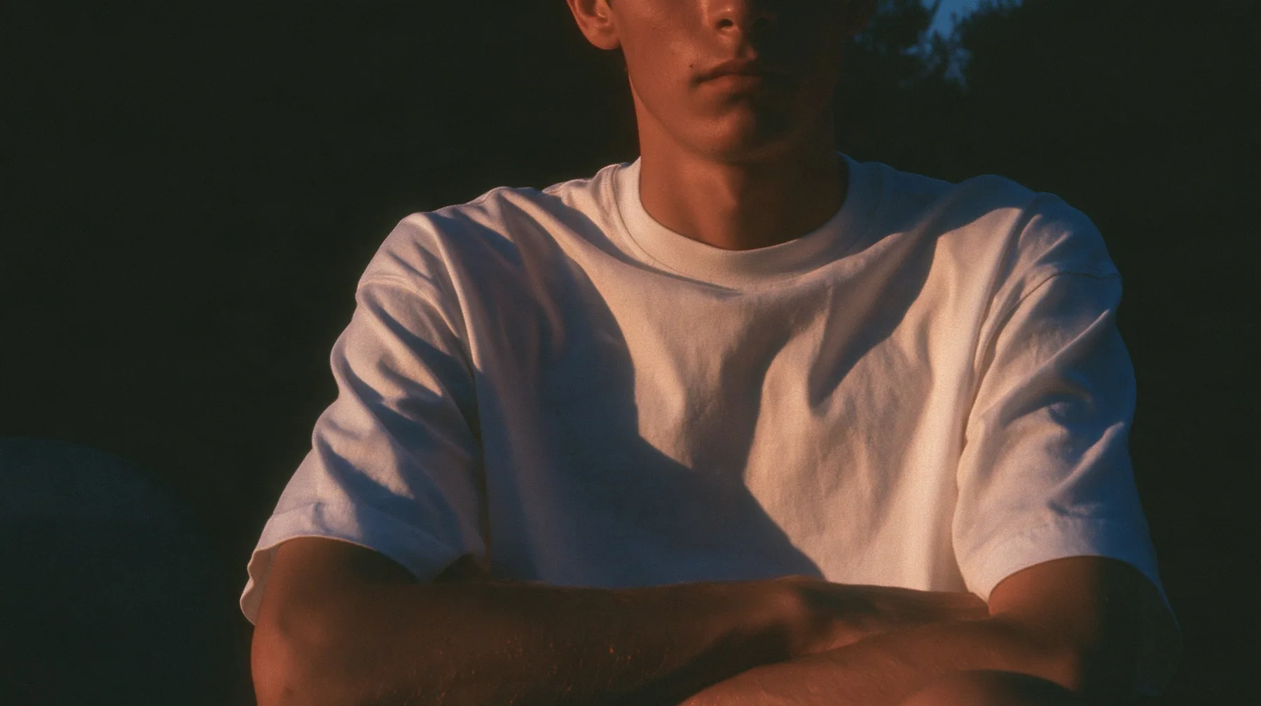 a front-row spectator in a plain white t-shirt seated in the dark, arms folded, face cropped at the chin, warm light, blue-black night turning to first light, clay-orange #D97757 light as the only colour, fine grain,…