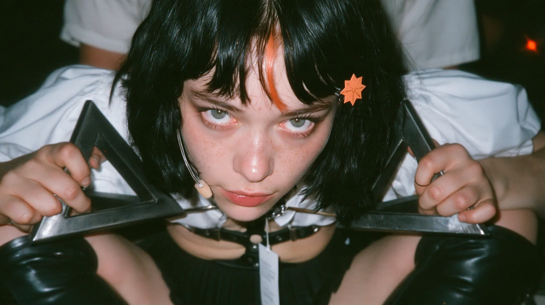 close-up of the hands of a young Caucasian American woman with pale skin and light freckles, a glossy black blunt jaw-length bob with heavy bangs and one clay-orange streak through the bangs, a small flat clay-orange…