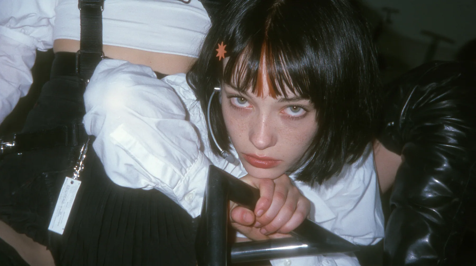 close-up of the hands of a young Caucasian American woman with pale skin and light freckles, a glossy black blunt jaw-length bob with heavy bangs and one clay-orange streak through the bangs, a small flat clay-orange…
