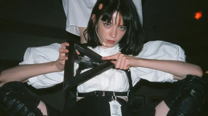 close-up of the hands of a young Caucasian American woman with pale skin and light freckles, a glossy black blunt jaw-length bob with heavy bangs and one clay-orange streak through the bangs, a small flat clay-orange…