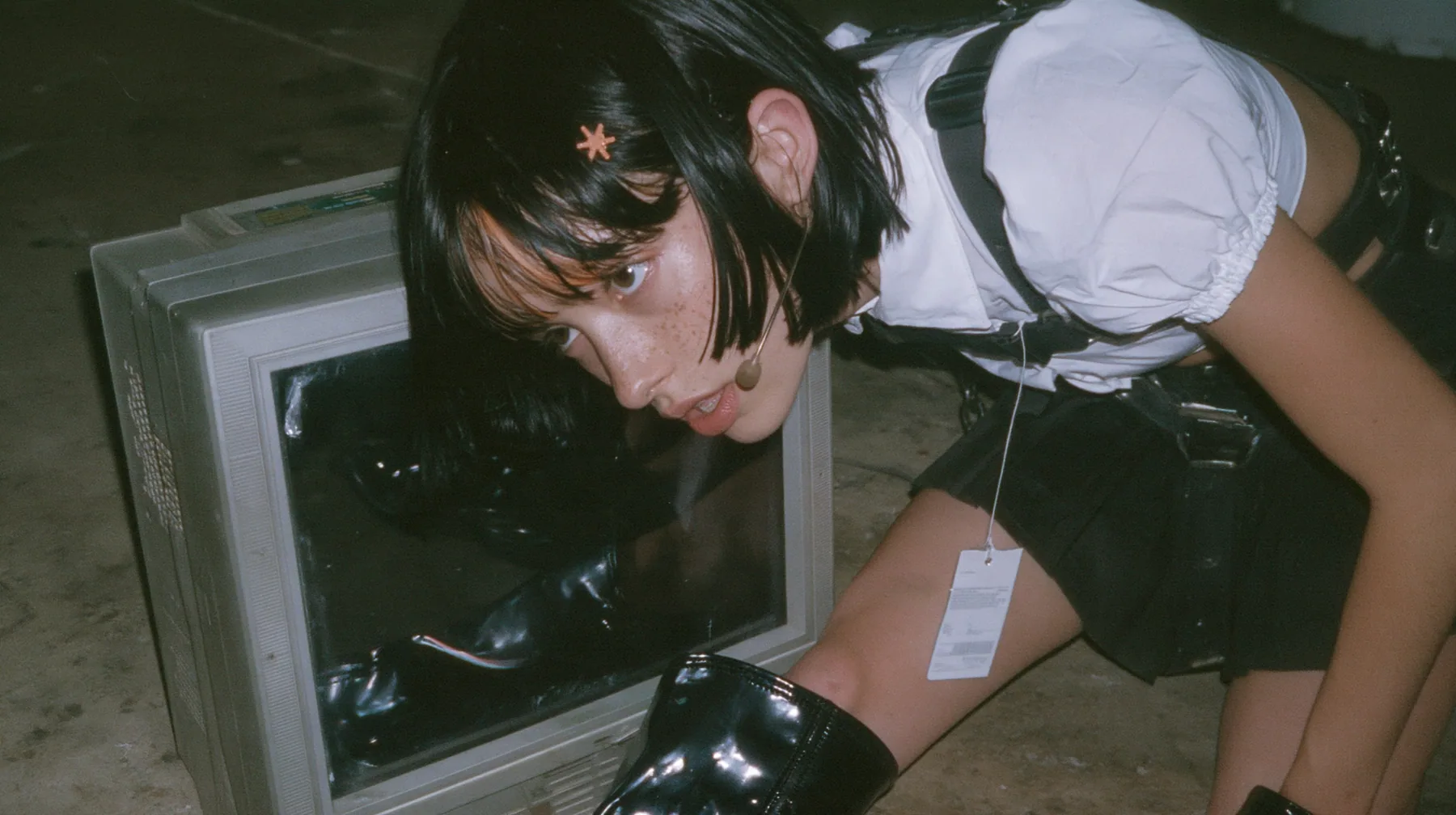 an old boxy monitor lying on a concrete floor with a black screen, the face of a young Caucasian American woman with pale skin and light freckles, a glossy black blunt jaw-length bob with heavy bangs and one…