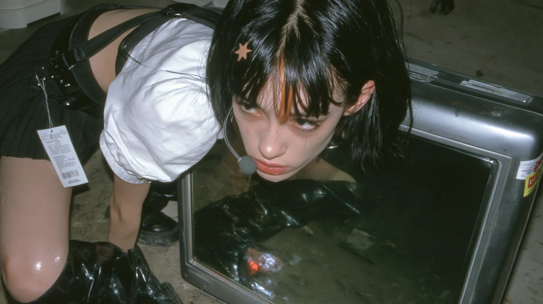 an old boxy monitor lying on a concrete floor with a black screen, the face of a young Caucasian American woman with pale skin and light freckles, a glossy black blunt jaw-length bob with heavy bangs and one…